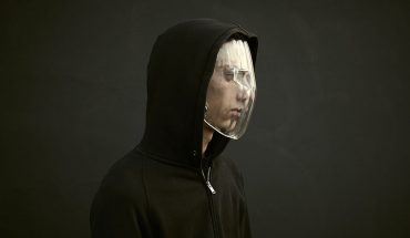 man in hoodie, looking off to right, wears transparent plastic face mask