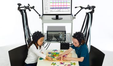 Two young children sit at table, playing with coloured objects. They wear 'skull caps', connected to wires leading to a computer-type device in the background.