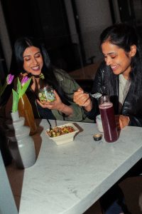two women smile and east as they sit at a table with fresh food offerings