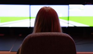 woman seated, looks ahead at three giant screens