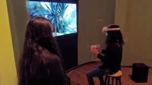 seated in front of video screen, a woman wears VR goggles and holds game controllers as another looks on