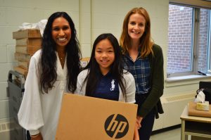 two women stand on either side of a young female student, who holds a HP computer box.