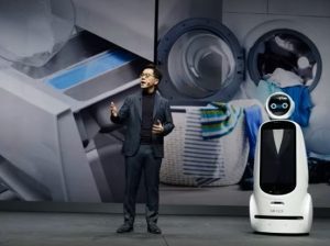 man speaks onstage accompanied by robot like device