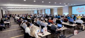 large conference room with attendees at desks, computer screens visible