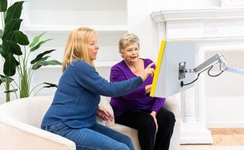 two women, seated, look at and point to computer touch screen on stand