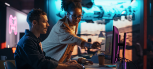 in workplace setting, a man sits at computer terminal; woman standing alongside points to screen