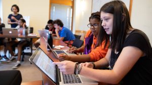 young students sit at computer laptops, discussing on screen content