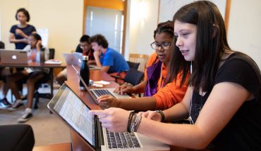 young students sit at computer laptops, discussing on screen content