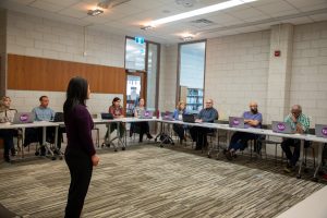 group of students seated around room listen to female instructor