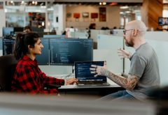 office workers seated in discussion, computers and desks in background