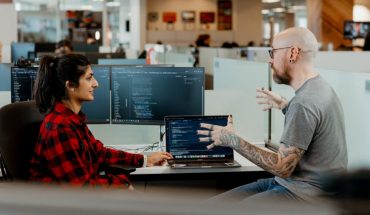 office workers seated in discussion, computers and desks in background