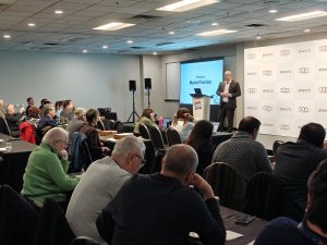 man on stage addresses seated crowd of attendees